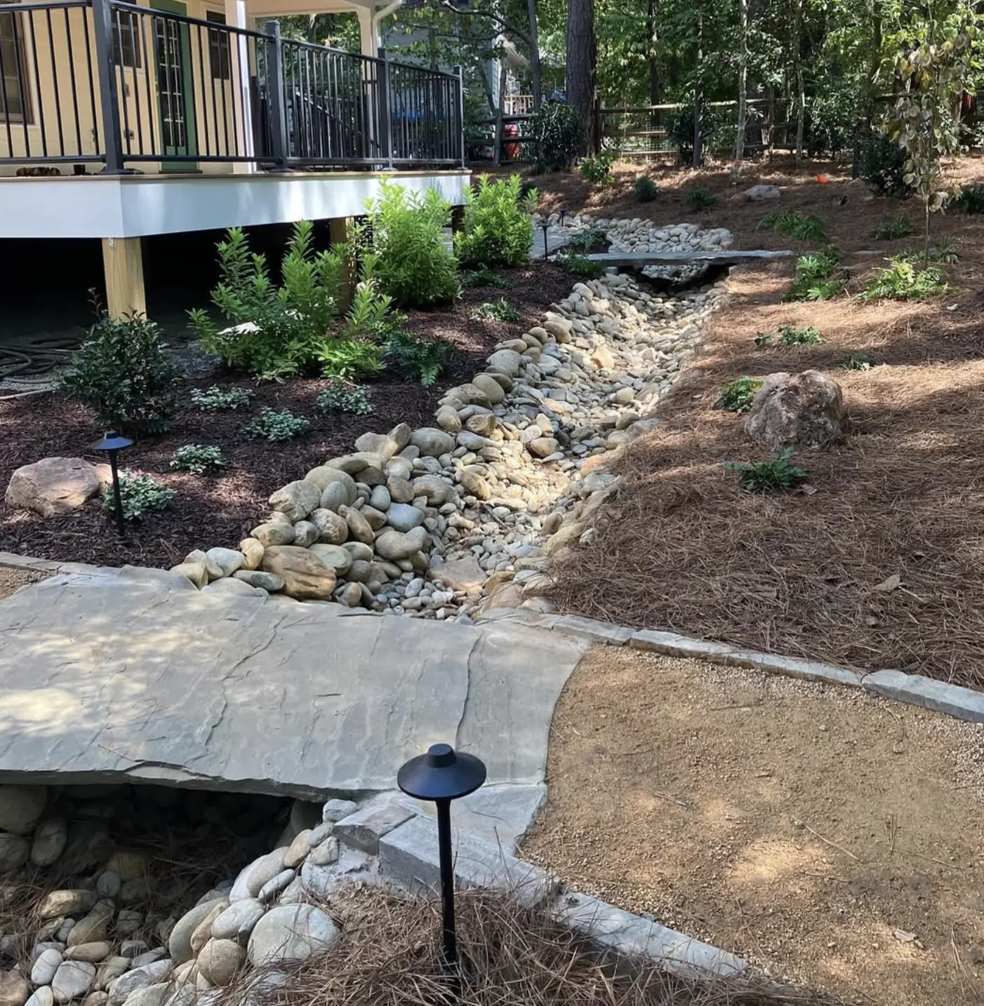 River rock dry creek bed carrying drainage past a raised deck with a flagstone crossing and path lights