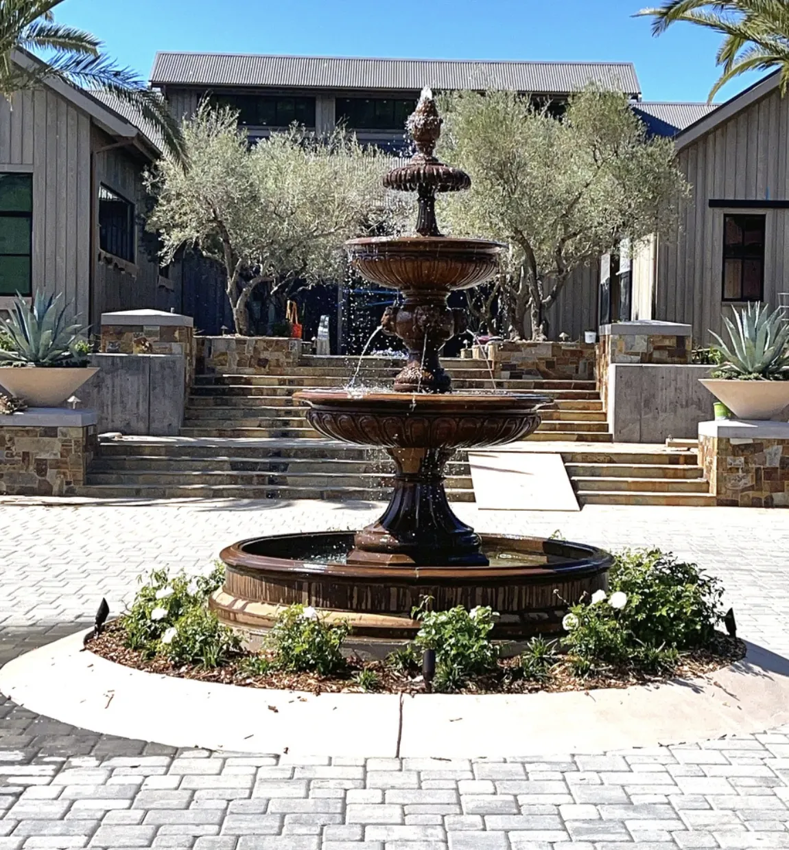 Dark bronze tiered fountain with a rose ring in a brick courtyard with olive trees