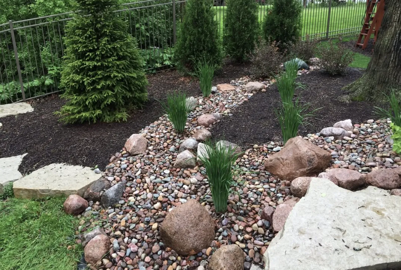Dry creek bed of mixed river rock with boulders and young grasses along a metal fence