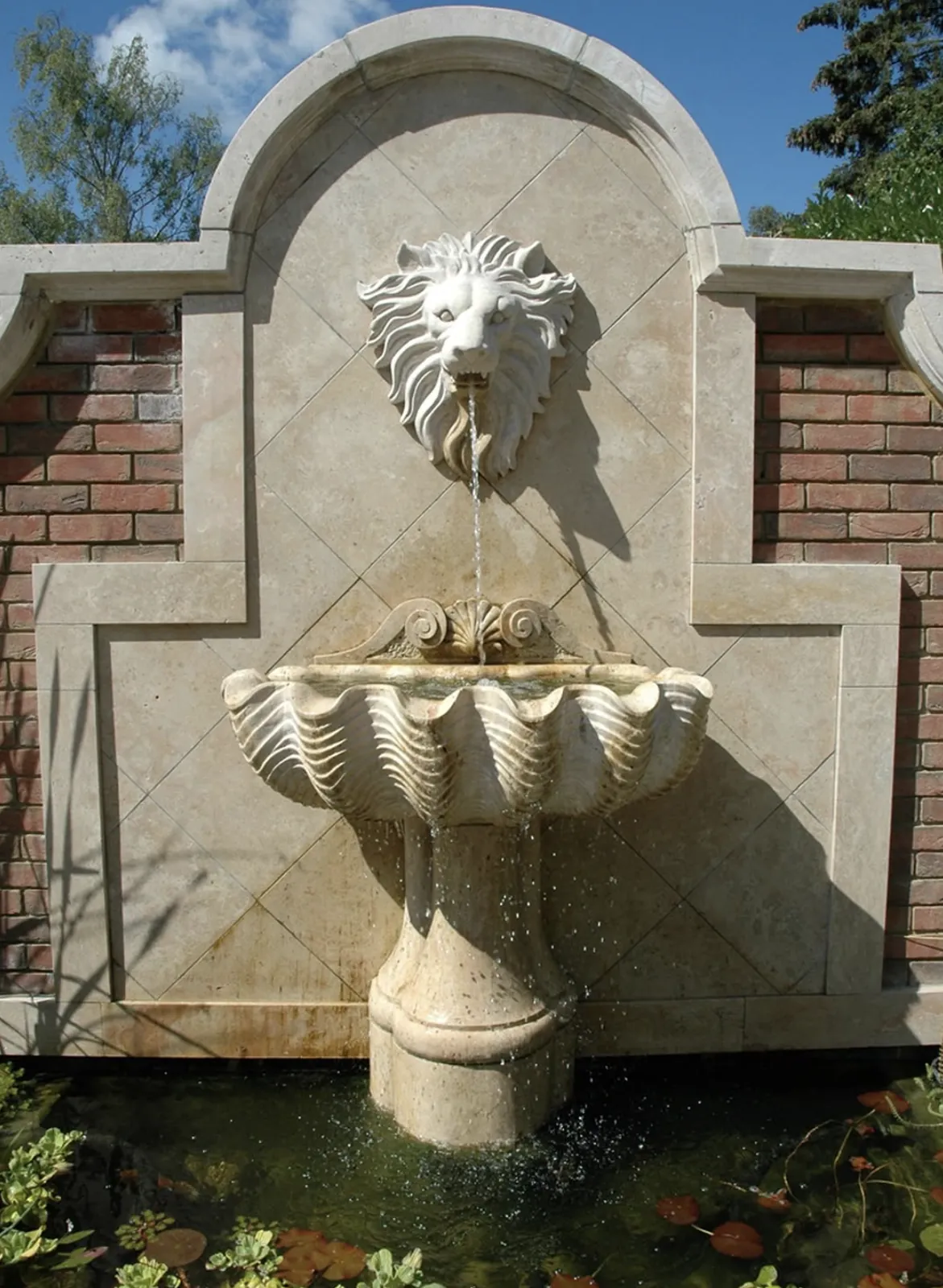 White marble lion head wall fountain with a shell-shaped basin trickling into a koi pond