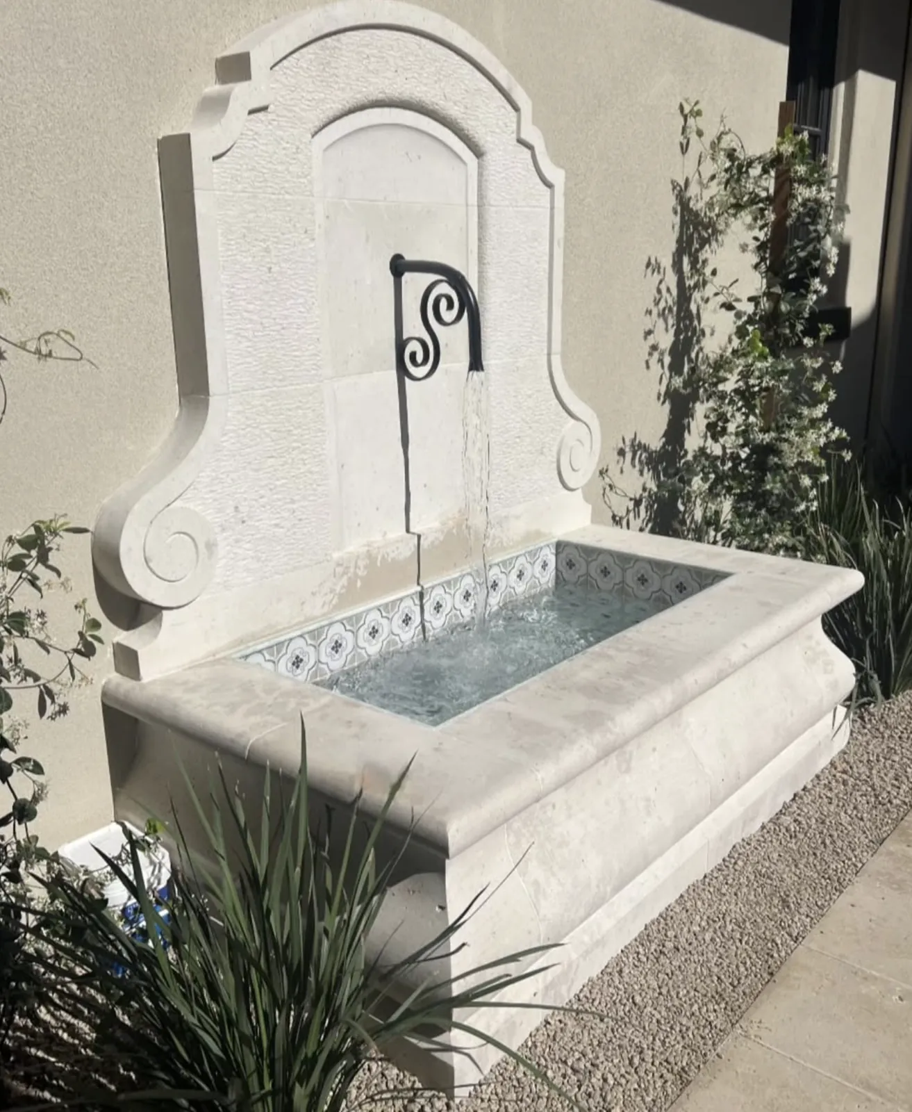Limestone wall fountain with a wrought iron spout pouring into a blue and white tiled trough