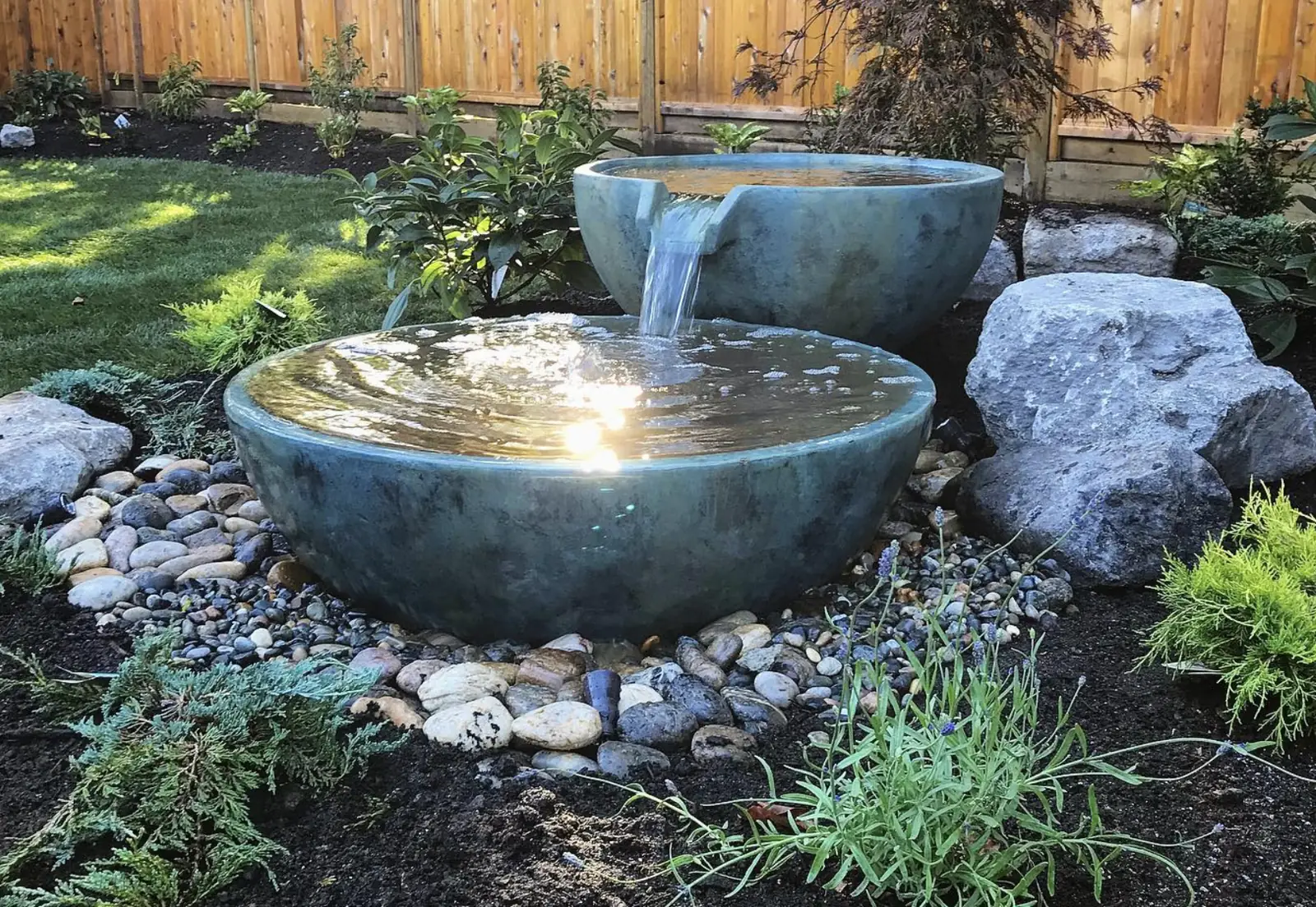 Two weathered green spillway bowls cascading water onto a river rock bed by a cedar fence